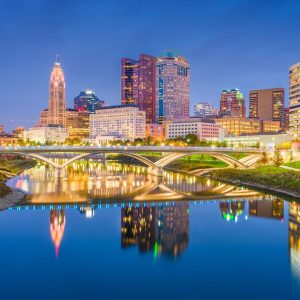 Twilight cityscape with illuminated buildings and reflective river.